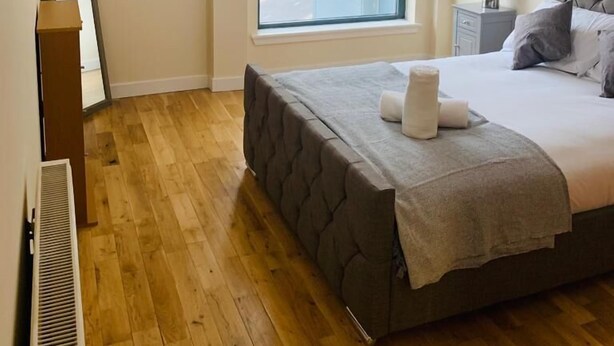 Un dormitorio con un ventanal grande, una cama con cabecera gris, una mesita de noche con lámpara y un radiador.