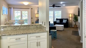 A modern kitchen with white cabinets and granite countertops, a dining area with a table and chairs, and a living room with a sofa and a coffee table. - OBX Sunset Lovers ! 2 Bed/2 Bath Soundside Condo with Canal View (Kill Devil Hills)