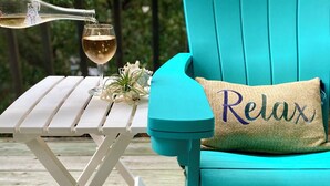 A turquoise Adirondack chair with a 'Relax' pillow, a white folding table, and a bottle of wine on a wooden deck. - OBX Sunset Lovers ! 2 Bed/2 Bath Soundside Condo with Canal View (Kill Devil Hills)