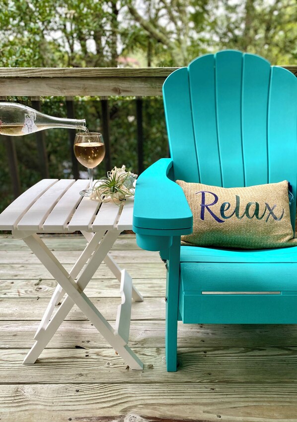 A turquoise Adirondack chair with a 'Relax' pillow, a white folding table, and a bottle of wine on a wooden deck. - OBX Sunset Lovers ! 2 Bed/2 Bath Soundside Condo with Canal View (Kill Devil Hills)