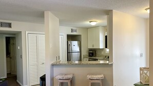 A compact kitchen with white cabinets, a stainless steel refrigerator, and a coffee maker. There are two stools and a small table. - OBX Sunset Lovers ! 2 Bed/2 Bath Soundside Condo with Canal View (Kill Devil Hills)