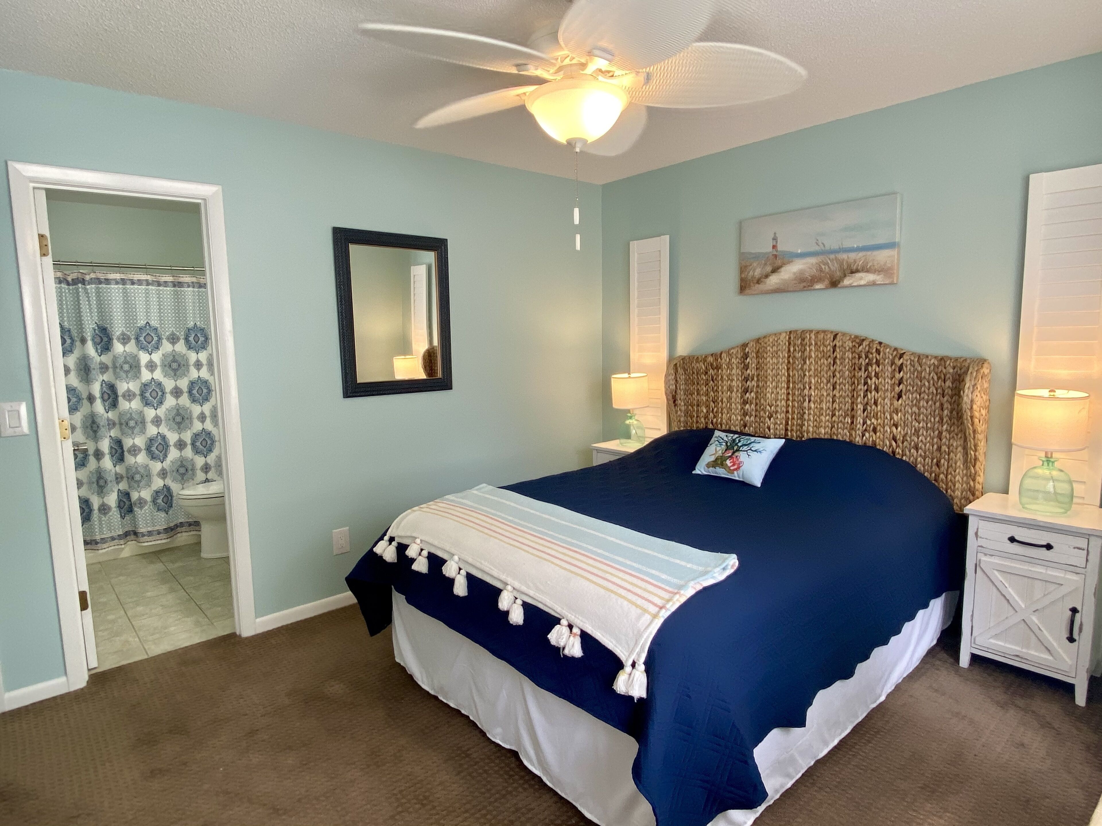 A bedroom with a bed, a ceiling fan, a nightstand with a lamp, a mirror, and a painting on the wall.