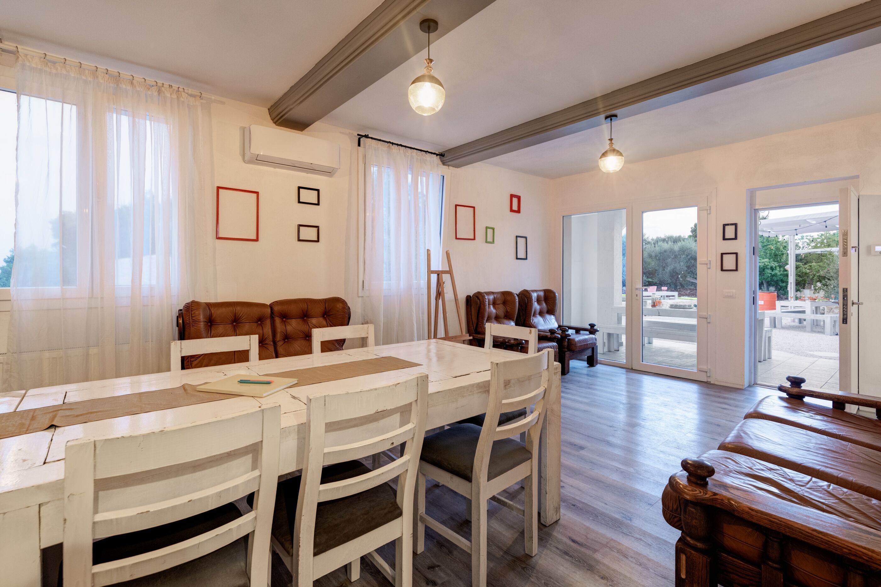 Un comedor con una mesa y sillas blancas, un banco de madera y vistas al exterior a través de amplios ventanales.