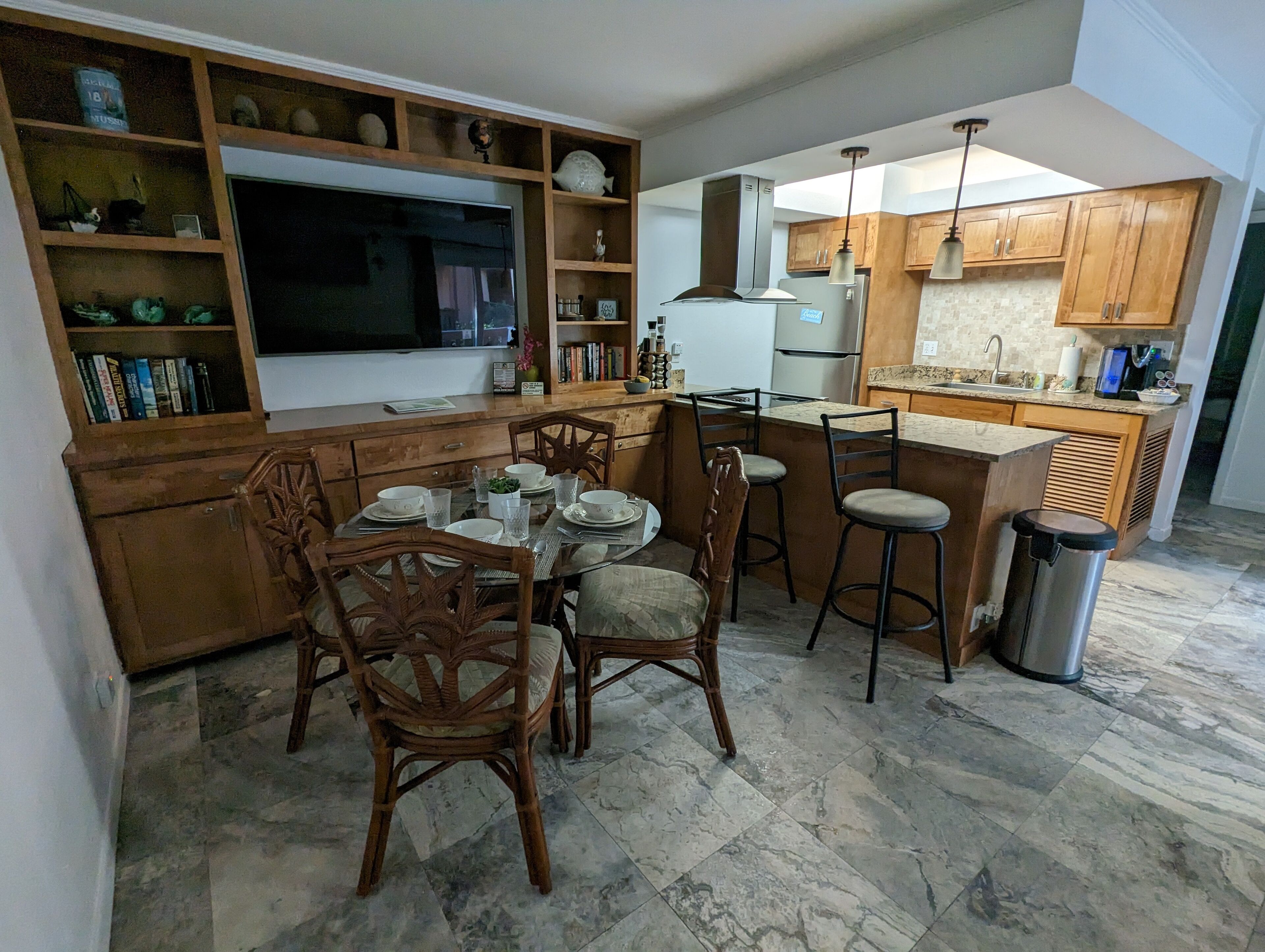A modern kitchen with wooden cabinets, a central island with stools, a built-in TV, and a dining area with chairs and a table set for four.