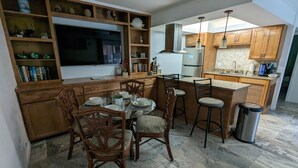 A modern kitchen with wooden cabinets, a central island with stools, a built-in TV, and a dining area with chairs and a table set for four. - Beachfront Condominium with Private Beach Access and Heated Pool (South Padre Island)