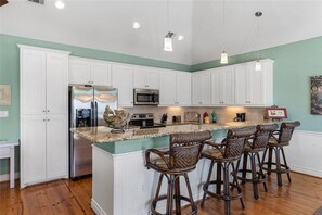 Una cocina con gabinetes blancos, un refrigerador de acero inoxidable, un microondas, un fregadero y una zona de bar con taburetes de mimbre.
