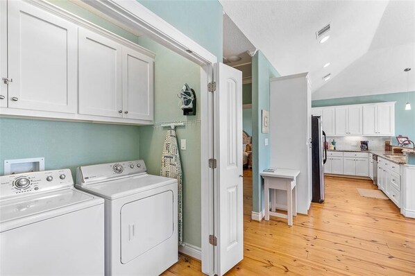 Un cuarto de lavado con lavadoras y secadoras apiladas, una mesita pequeña y una puerta que da a una zona de cocina.
