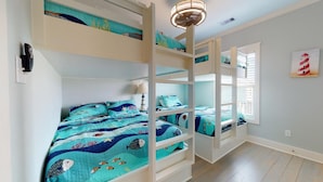 Une chambre avec un lit superposé, une couvre-lit aux motifs bleus évoquant l’océan, un ventilateur de plafond et une fenêtre avec des stores.