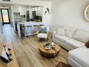 A modern living room with a white sofa, a wooden coffee table, and a kitchen area with white cabinets and a stainless steel refrigerator.