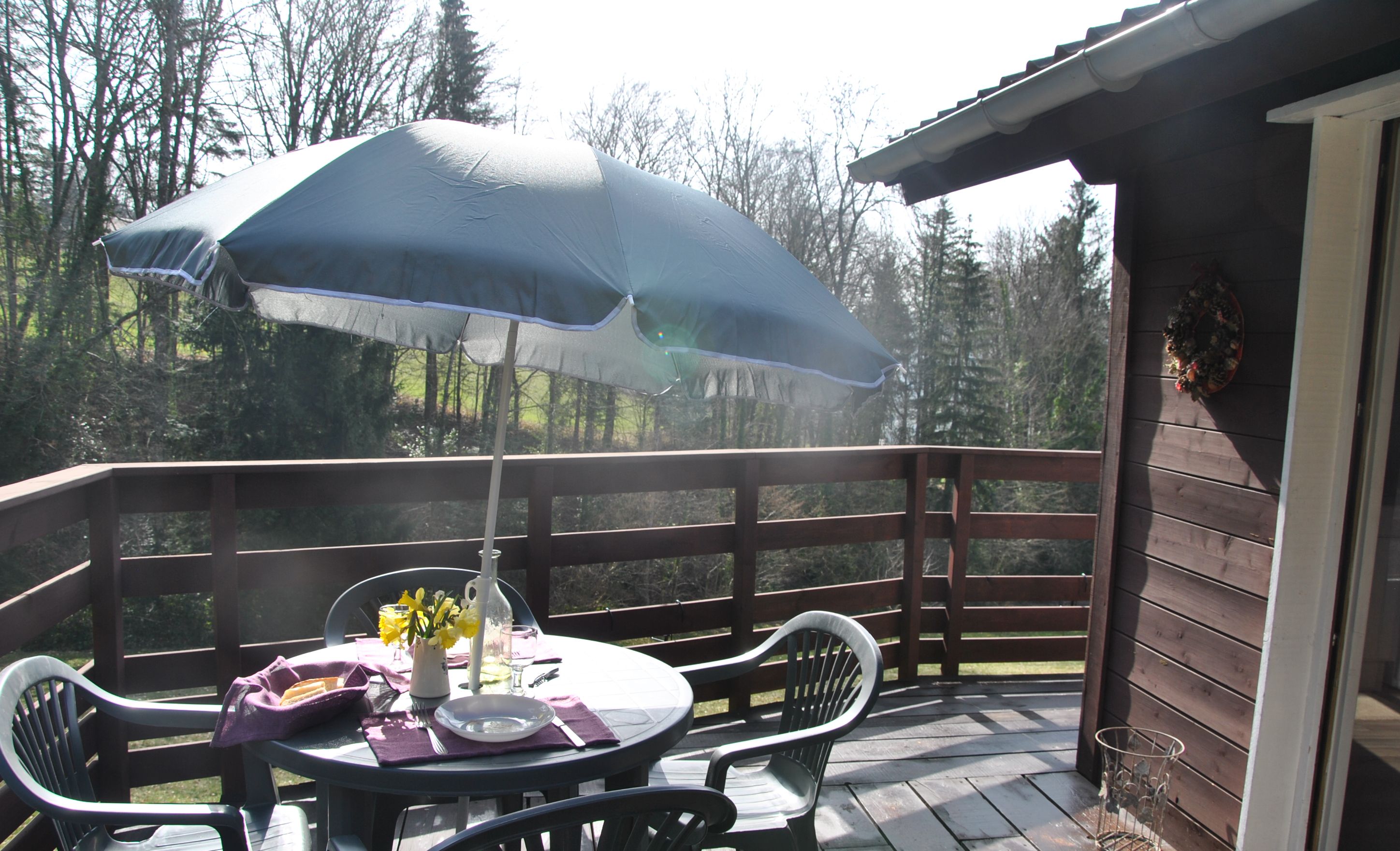 Une terrasse en bois avec une table et des chaises, un grand parasol et un vase rempli de fleurs.