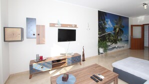 Una sala de estar con un televisor de pantalla plana montado en la pared, un mueble de madera para entretenimiento, una mesa de centro y un mural grande con motivos playeros.