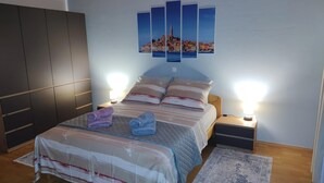 Un dormitorio con cama, dos mesitas de noche, un tocador, televisión montada en la pared y un ventanal grande.