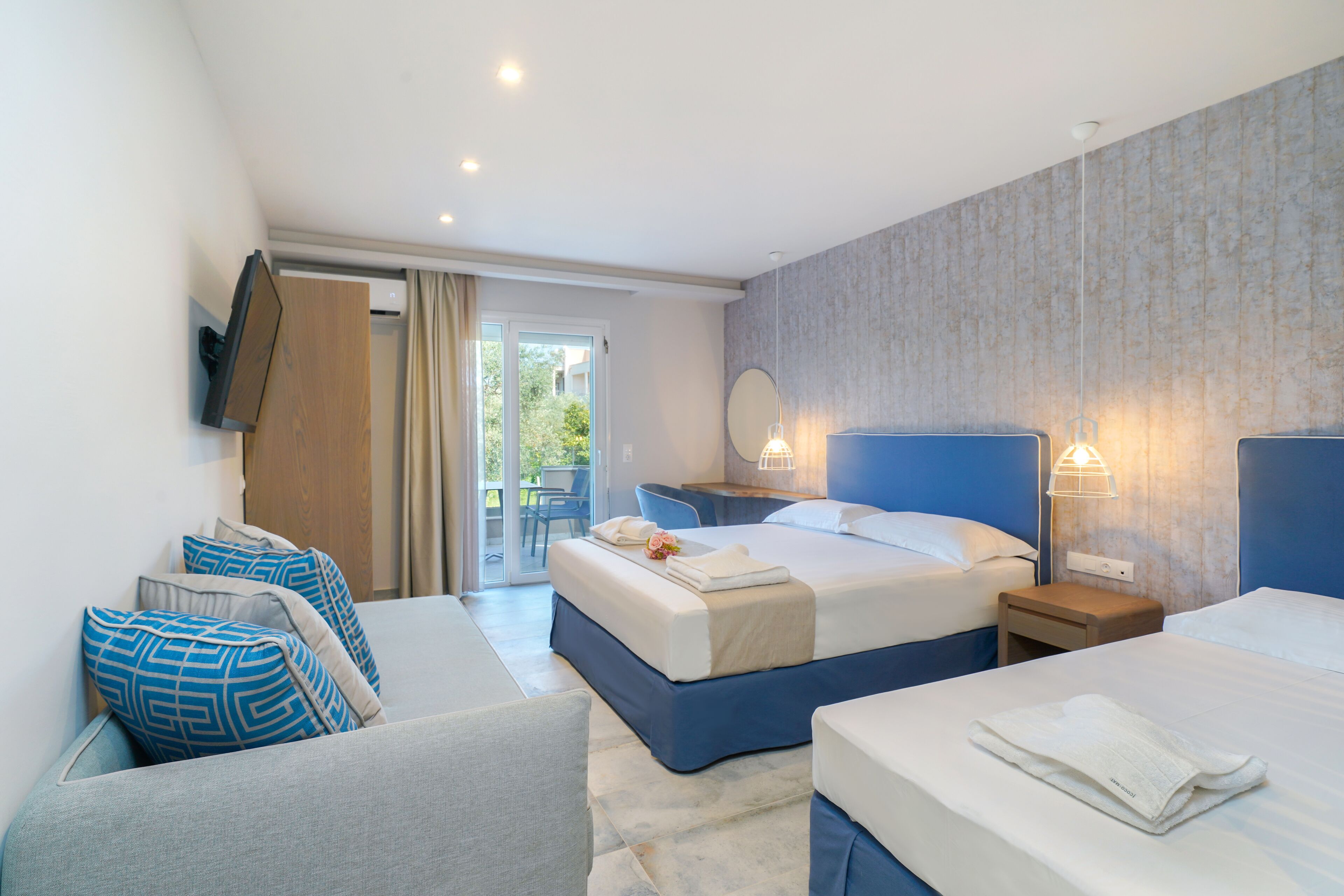 Hotelkamer met twee bedden, een bank, een klein tafeltje en een stoel. Er is een schuifdeur naar een buitengebied met een tafel en stoelen.