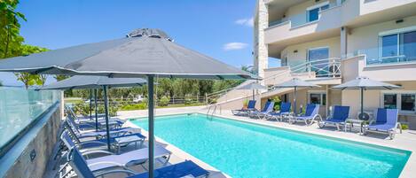 Una piscina con sillones de descanso y sombrillas, rodeada por un edificio con balcones.