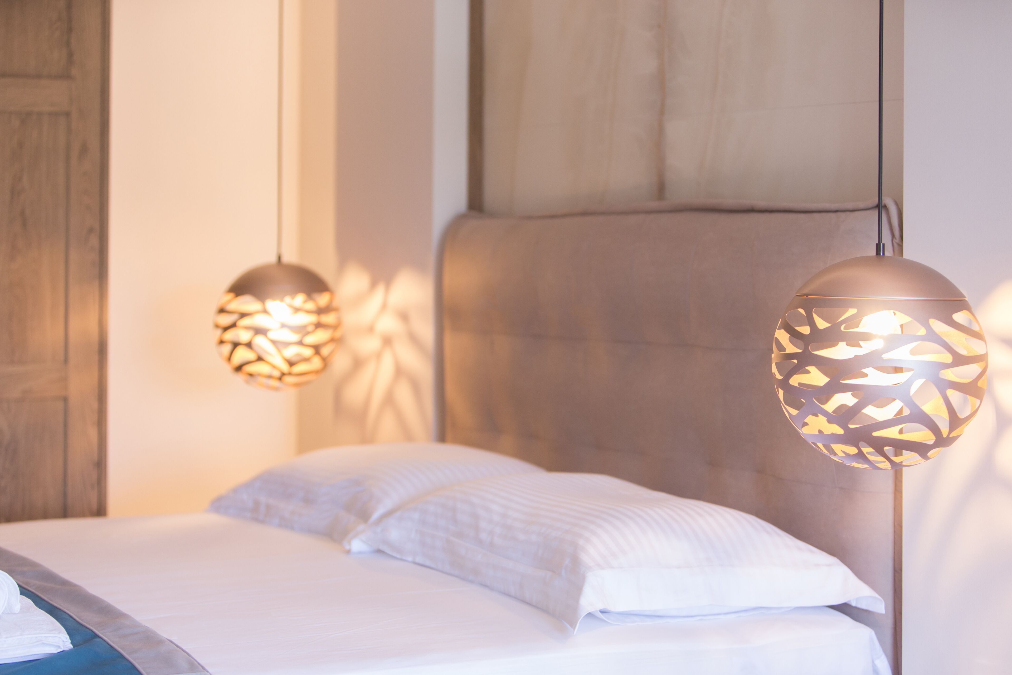 Ein Bett mit weißen Leinen und einem Kopfteil, zwei Pendelleuchten mit geometrischem Muster und eine Holztür im Hintergrund.