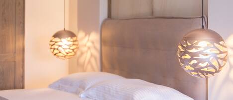 Ein Bett mit weißen Leinen und einem Kopfteil, zwei Pendelleuchten mit geometrischem Muster und eine Holztür im Hintergrund.