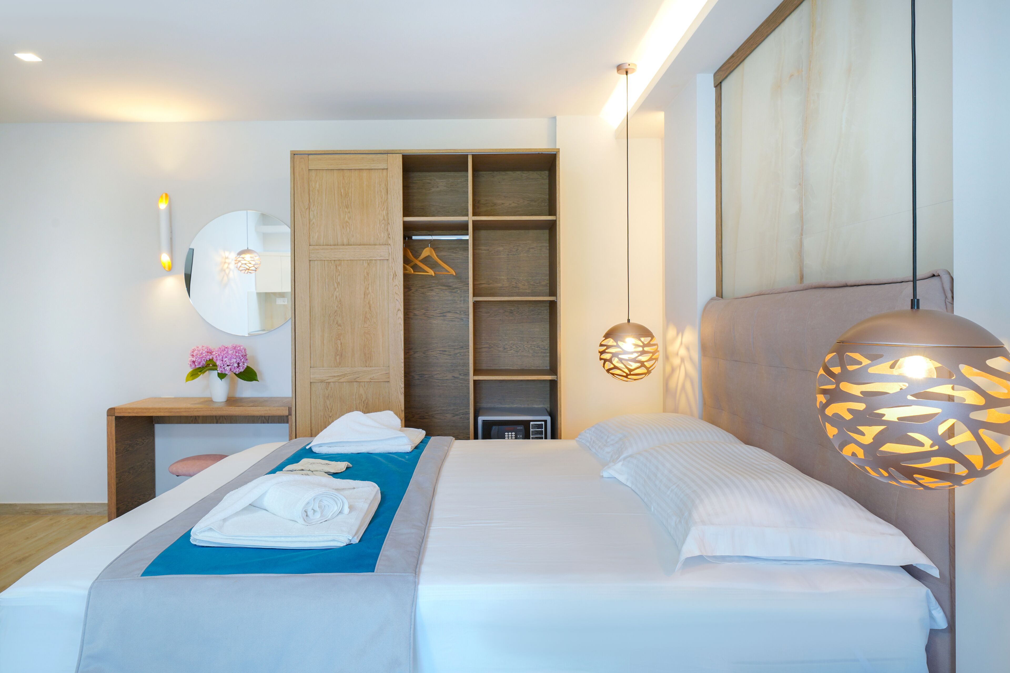 A modern hotel room with a large bed, a wardrobe, a bedside table with a lamp, and a mirror.