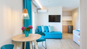 A modern living room with a blue sofa, teal chairs, a wooden dining table with a vase of flowers, and a kitchen area in the background.