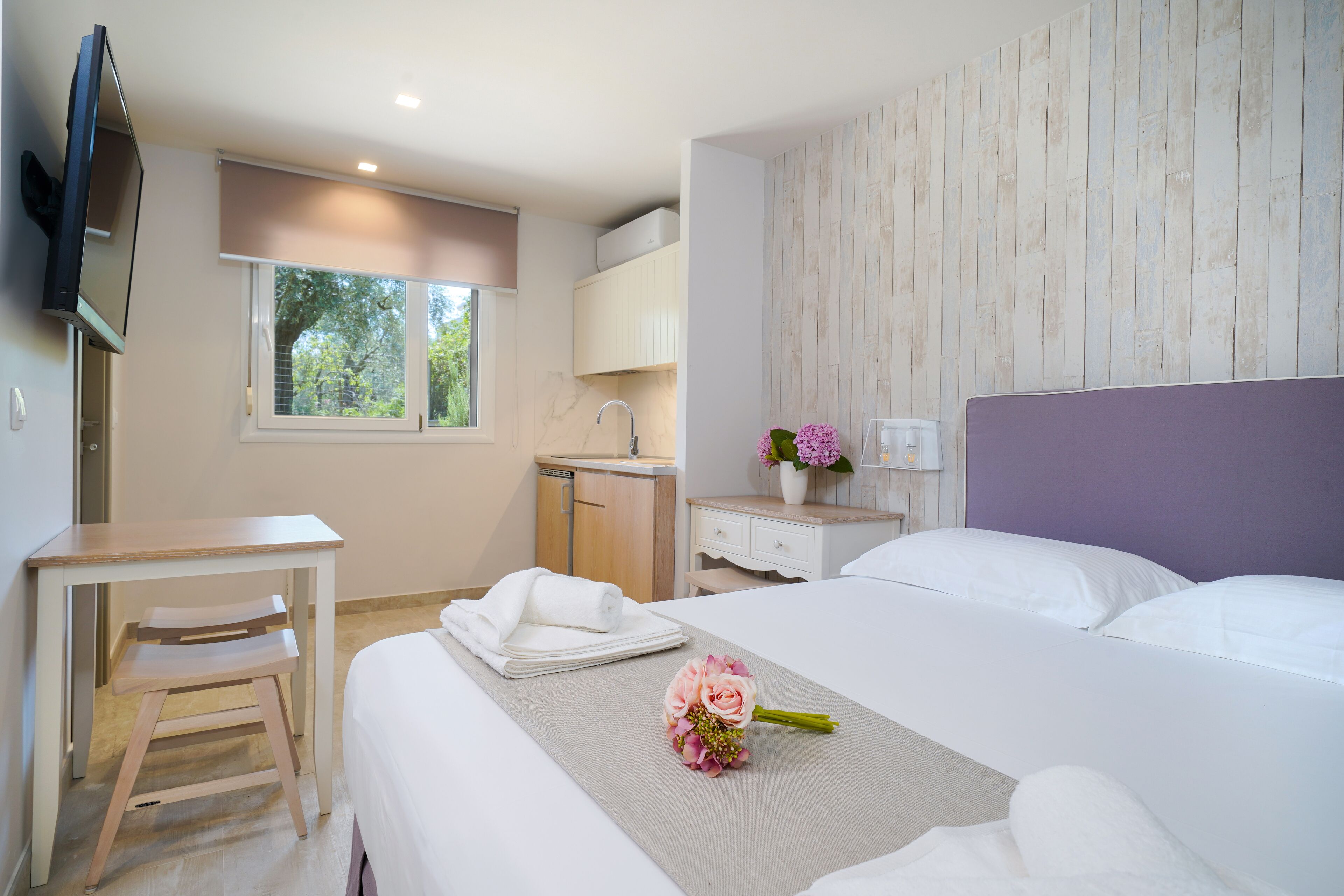 A hotel room with a bed, a desk, a chair, a TV, a window, and a wooden wall panel.
