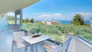 A balcony with a white table and chairs, a potted plant, and a view of a coastal area with buildings and greenery.