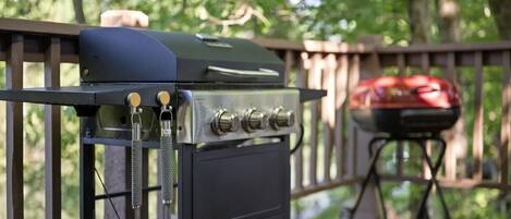 Un barbecue sur une terrasse en bois, avec un panier à griller et une housse de protection pour le barbecue en arrière-plan.