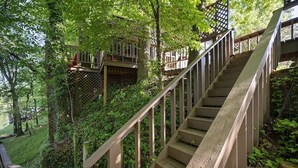 Eine Holztreppe mit Geländer führt zu einer hölzernen Terrasse, die von üppigem Grün umgeben ist.