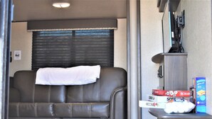 A compact RV interior with a leather sofa, a window with blinds, and a small kitchenette area with a microwave and a box of cereal. - Grand Canyon RV Glamping Luxury Suite (Glamping Getaway) (Williams)