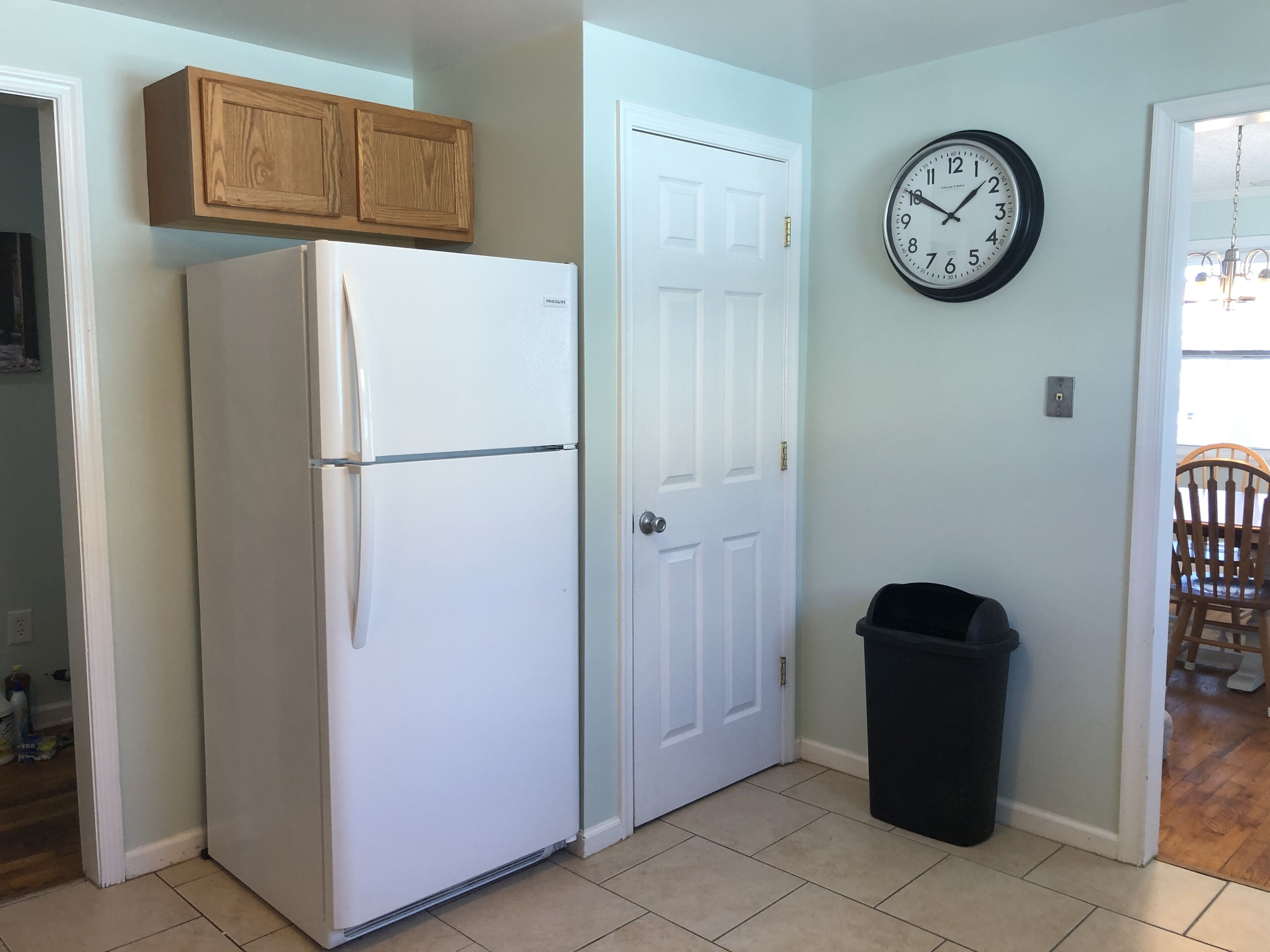 Una cocina con un refrigerador blanco, armarios de madera, una puerta blanca, un cubo de basura negro y un reloj de pared.