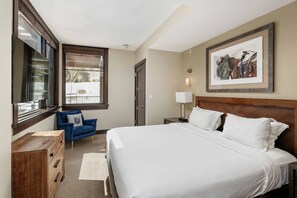 A hotel room with a large bed, a blue armchair, a wooden dresser, a window with blinds, and a framed picture on the wall. - Hayden Lodge by iTrip Aspen Snowmass (Snowmass Village)