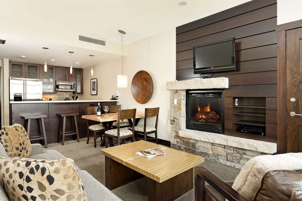 A modern living room with a stone fireplace, a flat-screen TV mounted above it, and a dining area with a kitchen in the background.
