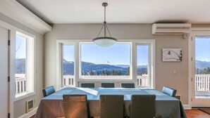 Een eetruimte met een grote tafel en stoelen, grote ramen met uitzicht op een berg, en een airconditioningapparaat aan de muur.