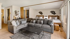 Uma sala de estar moderna com um sofá modular cinza, uma mesa de centro preta e piso de madeira. Há uma cama com cabeceira de madeira e um banquinho pequeno de madeira.