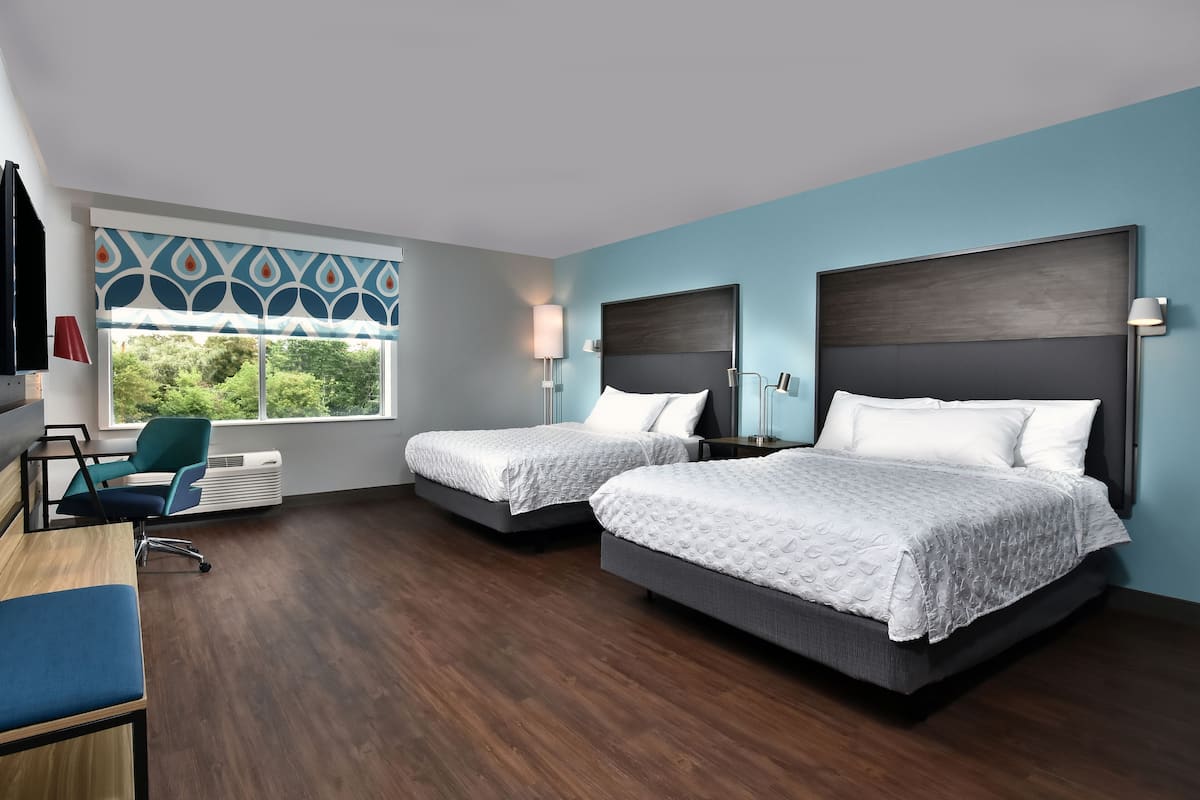 A hotel room with two beds, a desk, a chair, and a window with a view of greenery.