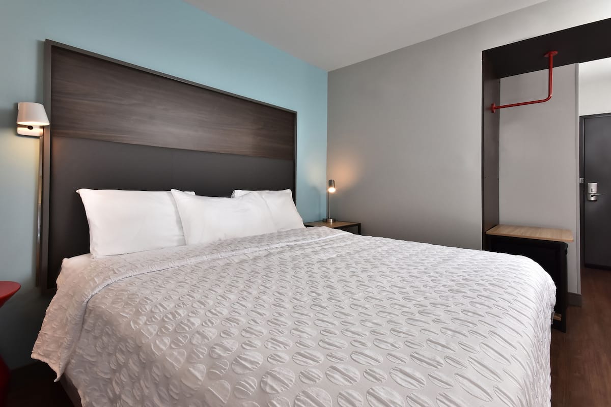A hotel room with a large bed, a nightstand, a wall-mounted lamp, and a door with a red handle.