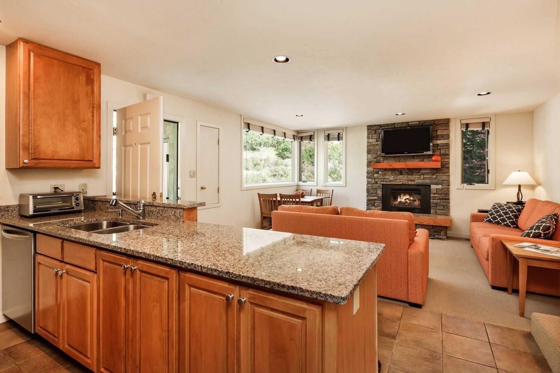 A kitchen with granite countertops, wooden cabinets, and a built-in oven. There is a dining area with a table and chairs, and a living room with a fireplace and seating.