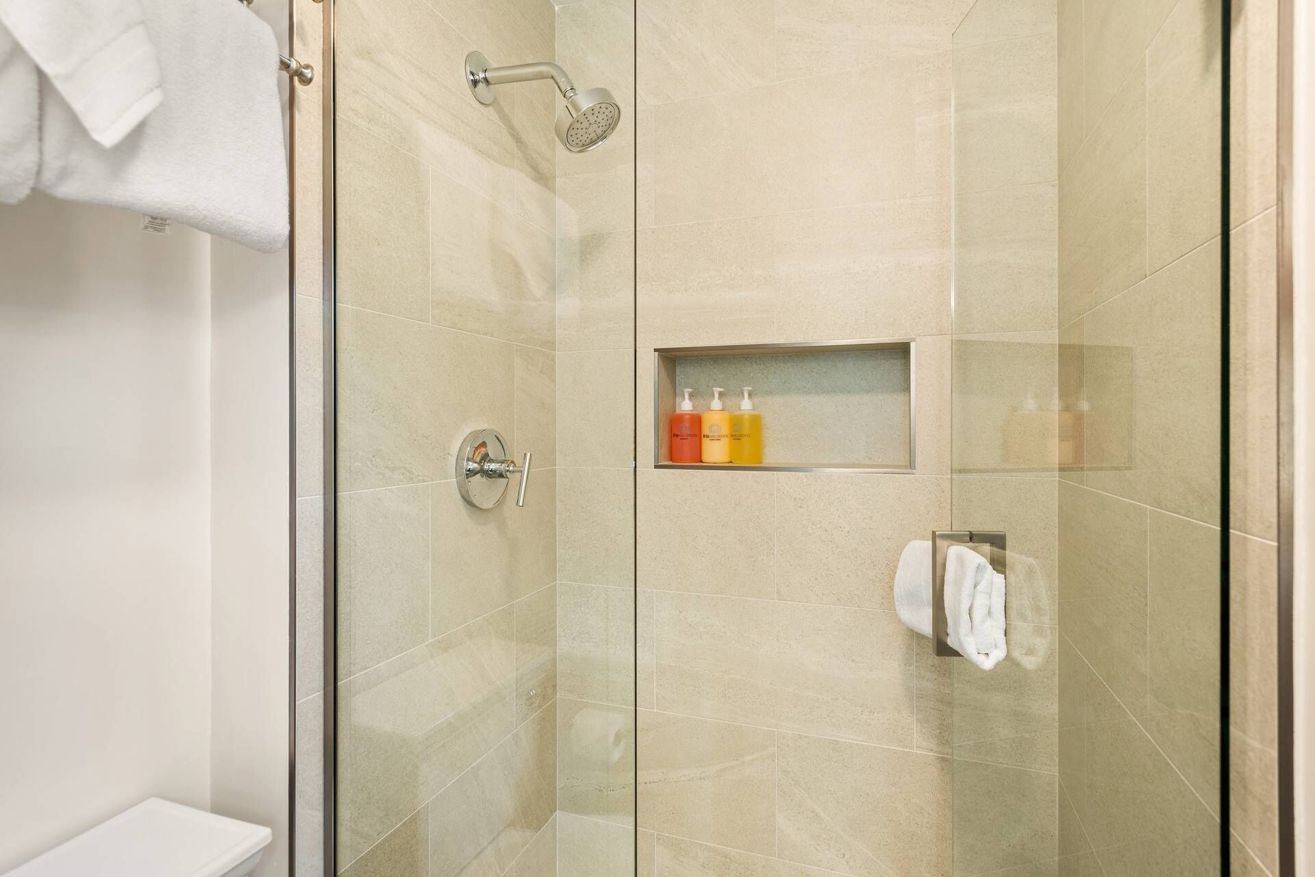 A glass-enclosed shower with a handheld showerhead, a shelf with toiletries, and a towel rack.