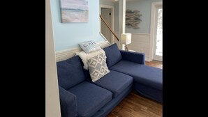 Ein Wohnzimmer mit einem blauen Ecksofa, einem Strandbild und einem Kissen mit der Aufschrift 'CAP HOUSE'.