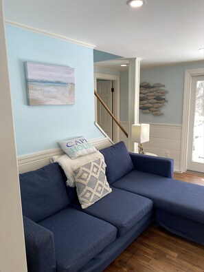 Un salon avec un canapé d’angle bleu, un tableau représentant une plage et un coussin orné de l’inscription « CAP HOUSE ».