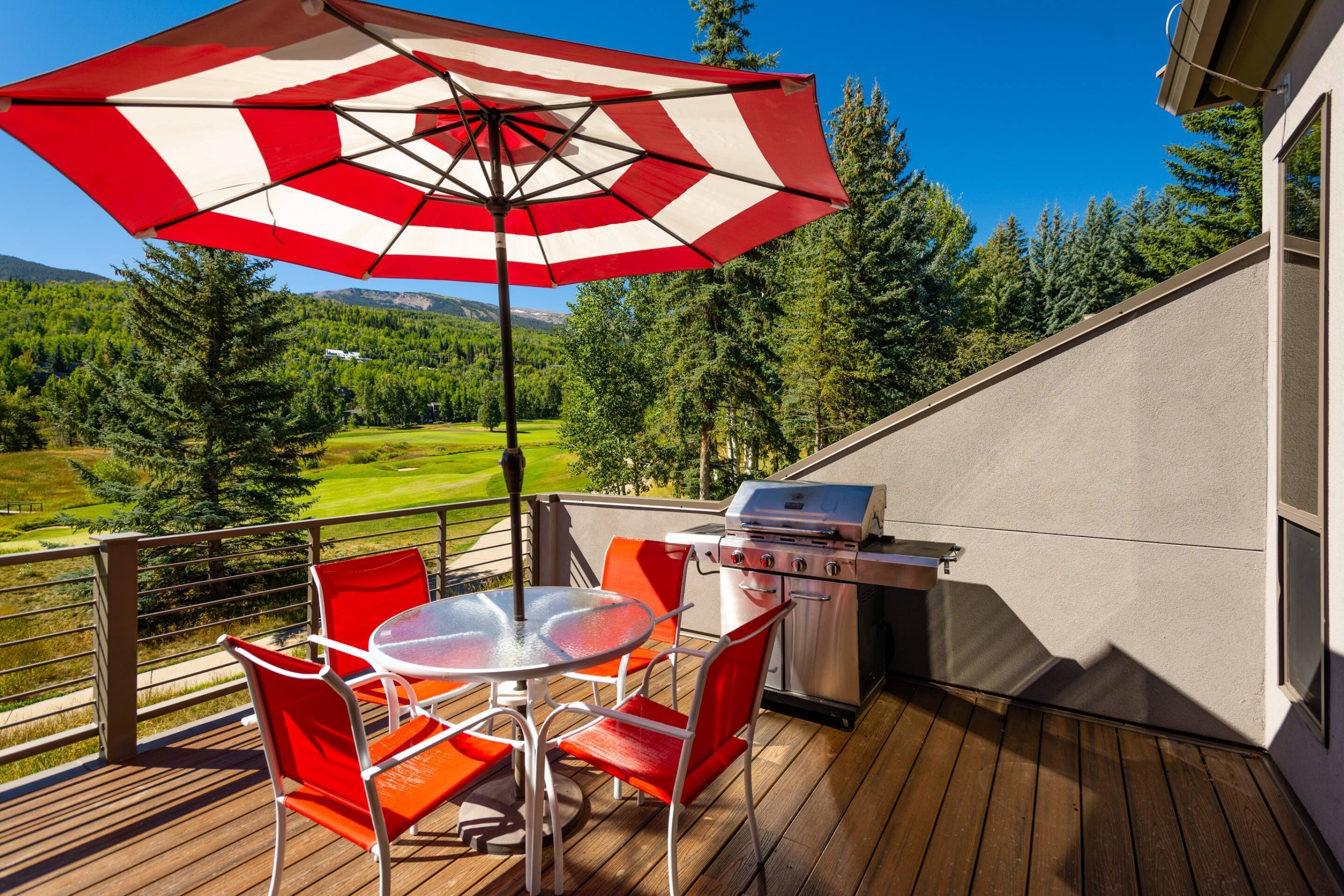 A deck with a grill, red chairs, and a table under a striped umbrella.