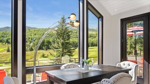 En matplats med ett stort bord och stolar, en modern lampa och utsikt över ett grönskande landskap genom stora fönster.