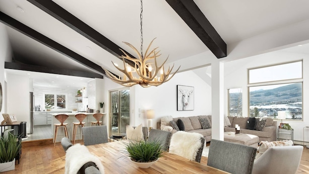 Un vaste salon doté d’une grande table à manger en bois, de chaises grises à revêtement en tissu et d’un plaid en fausse fourrure blanche. Un lustre orné de luminaires en forme de bois de cerf est suspendu au plafond.