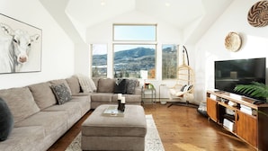Un salon spacieux avec un plafond haut, une grande fenêtre offrant une vue sur les montagnes, un canapé confortable, un meuble en bois avec un téléviseur et un fauteuil suspendu.