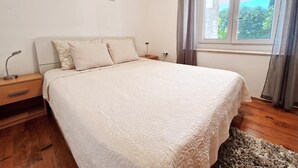 A bedroom with a bed, a nightstand, a lamp, a window with curtains, and a wall art piece. - Apartment Gina (Bale)