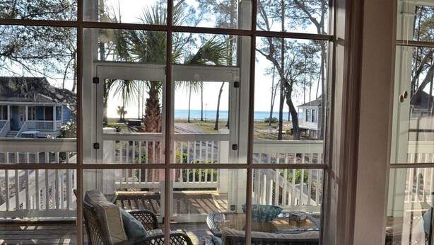 Blick durch eine Moskitonetz-Veranda mit Gartenmöbeln, einem weißen Zaun sowie auf den Strand und das Meer.