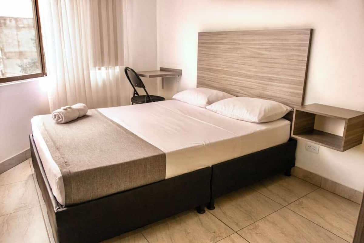 A neatly made bed with a headboard, a bedside table, a chair, and a small shelf unit.