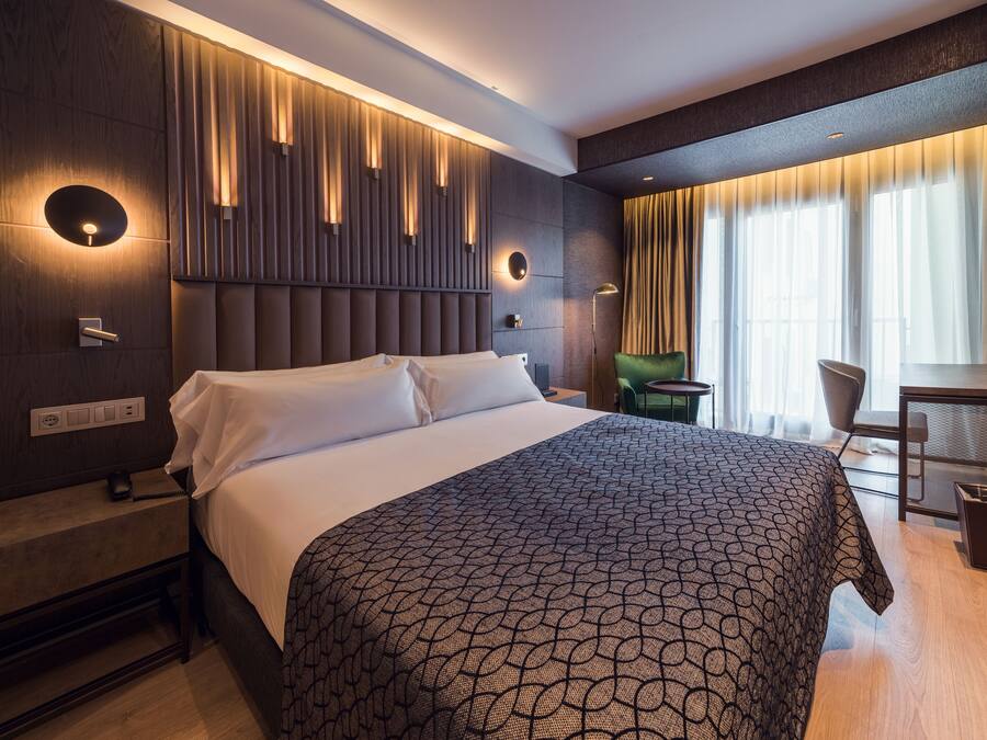A modern hotel room with a large bed, a desk, and a chair. There are wall-mounted lights and a window with curtains.