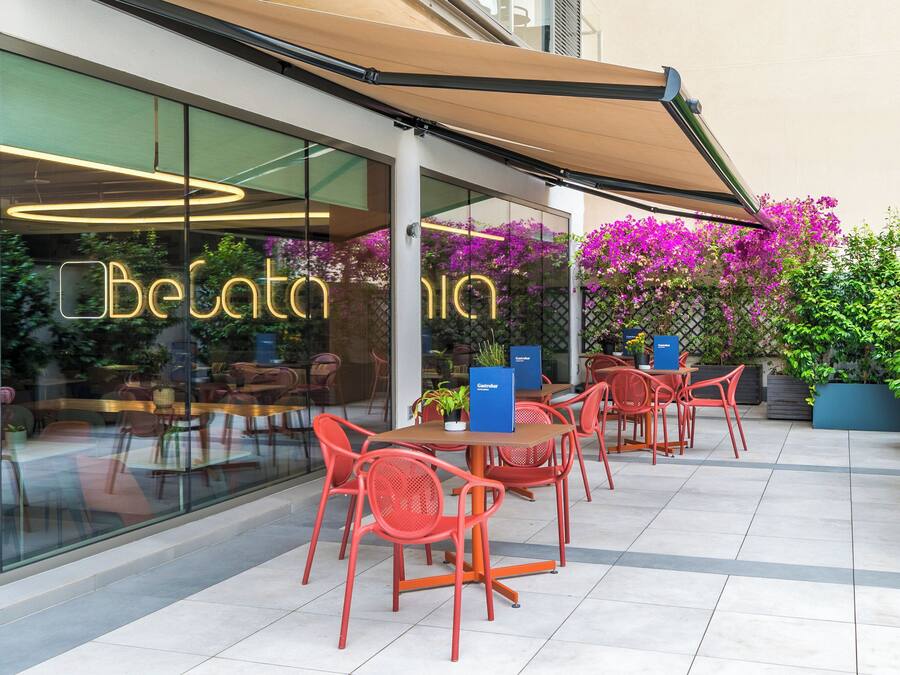 An outdoor seating area with red chairs and a table, a glass wall with the name 'BeGalleria' illuminated, and a potted plant with purple flowers.