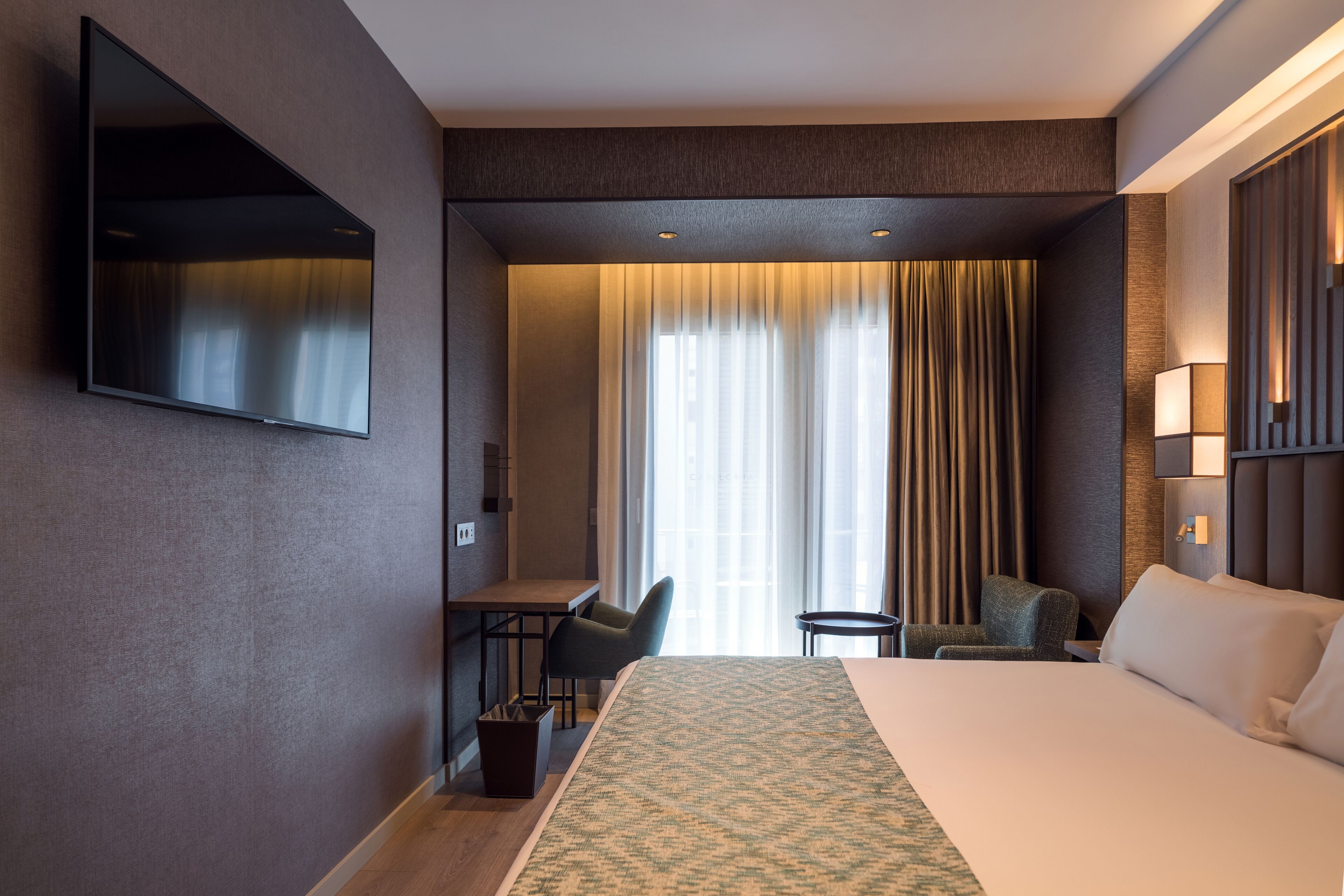 Habitación de hotel con una cama grande, un escritorio, una silla, un televisor montado en la pared y una ventana con cortinas.