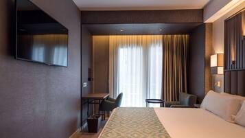 Habitación de hotel con una cama grande, un escritorio, una silla, un televisor montado en la pared y una ventana con cortinas.