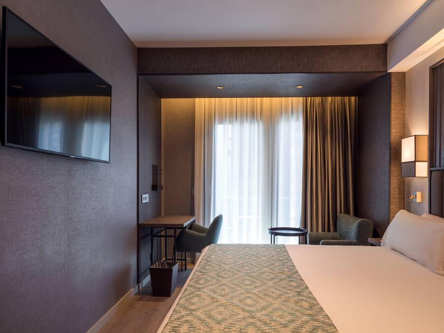 A hotel room with a large bed, a desk, a chair, a TV mounted on the wall, and a window with curtains.